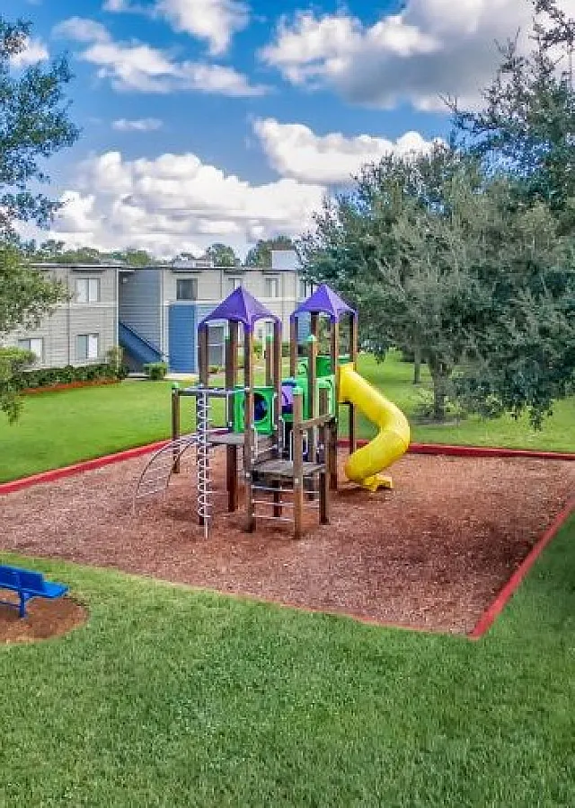 Charming outdoor playground surrounded by lush greenery and modern apartment buildings, featuring colorful slides and climbing structures.