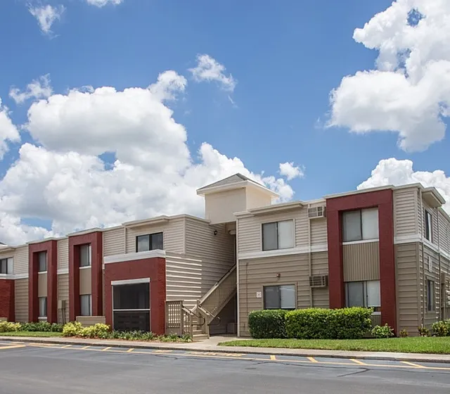 Charming modern apartment building with red and beige accents under a bright blue sky, surrounded by lush greenery and a peaceful neighborhood setting.