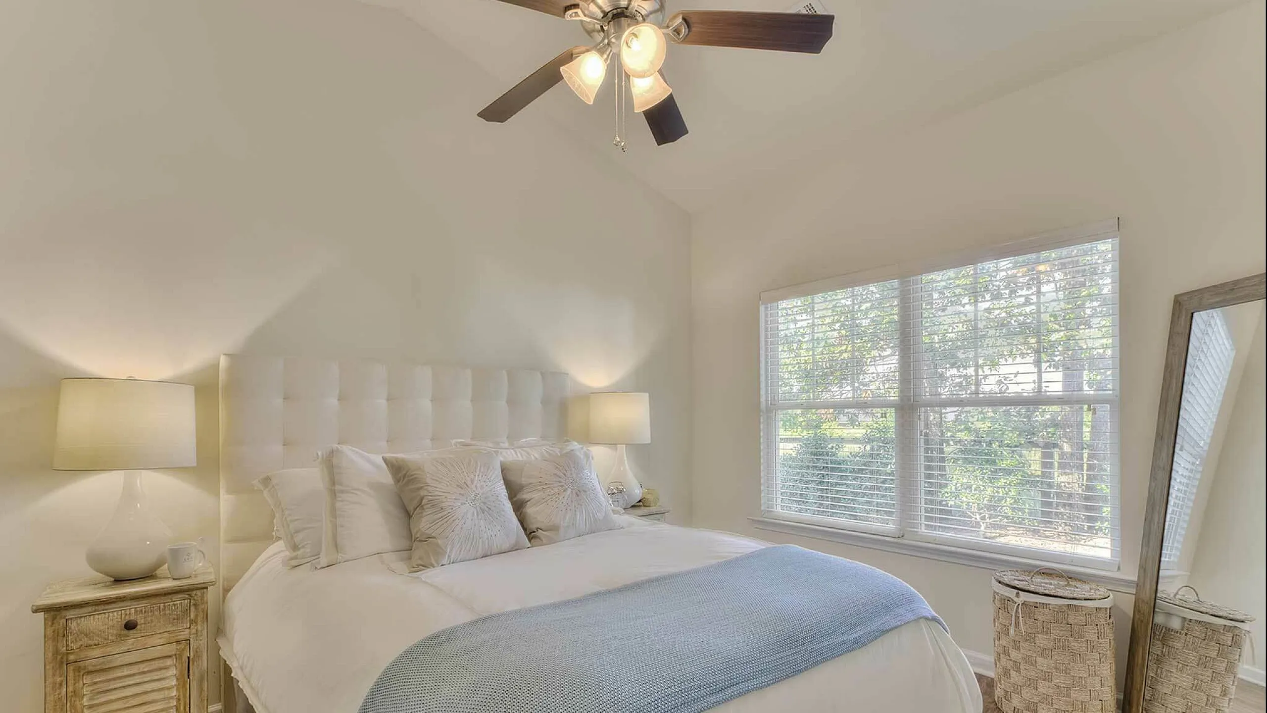 Bright and inviting bedroom with elegant decor, featuring a cozy bed adorned with plush pillows, a textured blue throw, and warm ambient lighting from matching lamps.