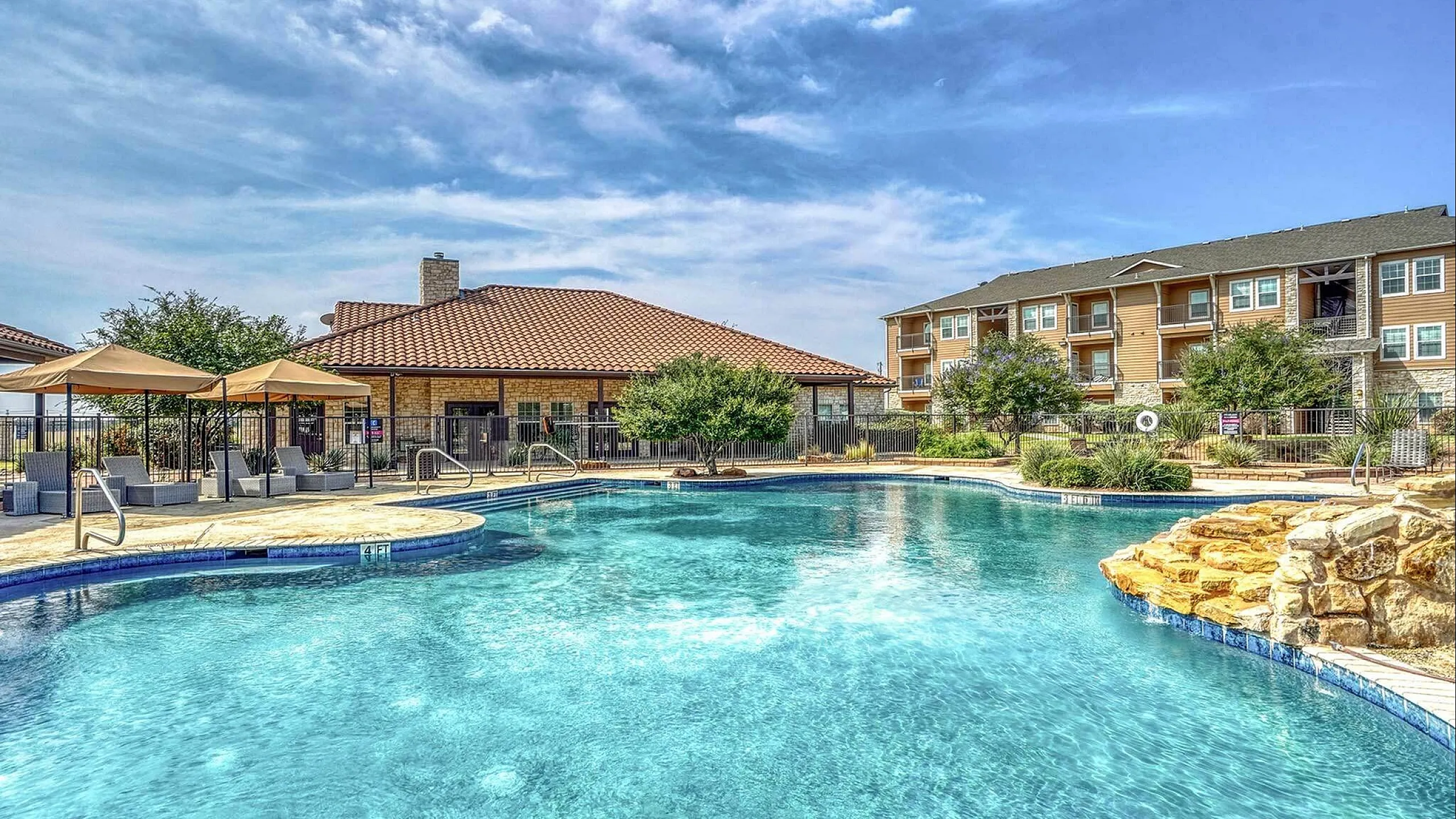Inviting apartment complex with a glistening outdoor pool, stylish poolside cabanas, and lush gardens under a clear, blue sky.