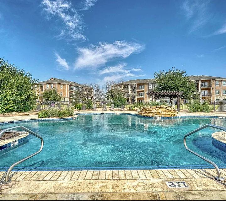 Inviting pool area with crystal-clear water surrounded by modern apartment buildings, lush greenery, and a stunning blue sky.