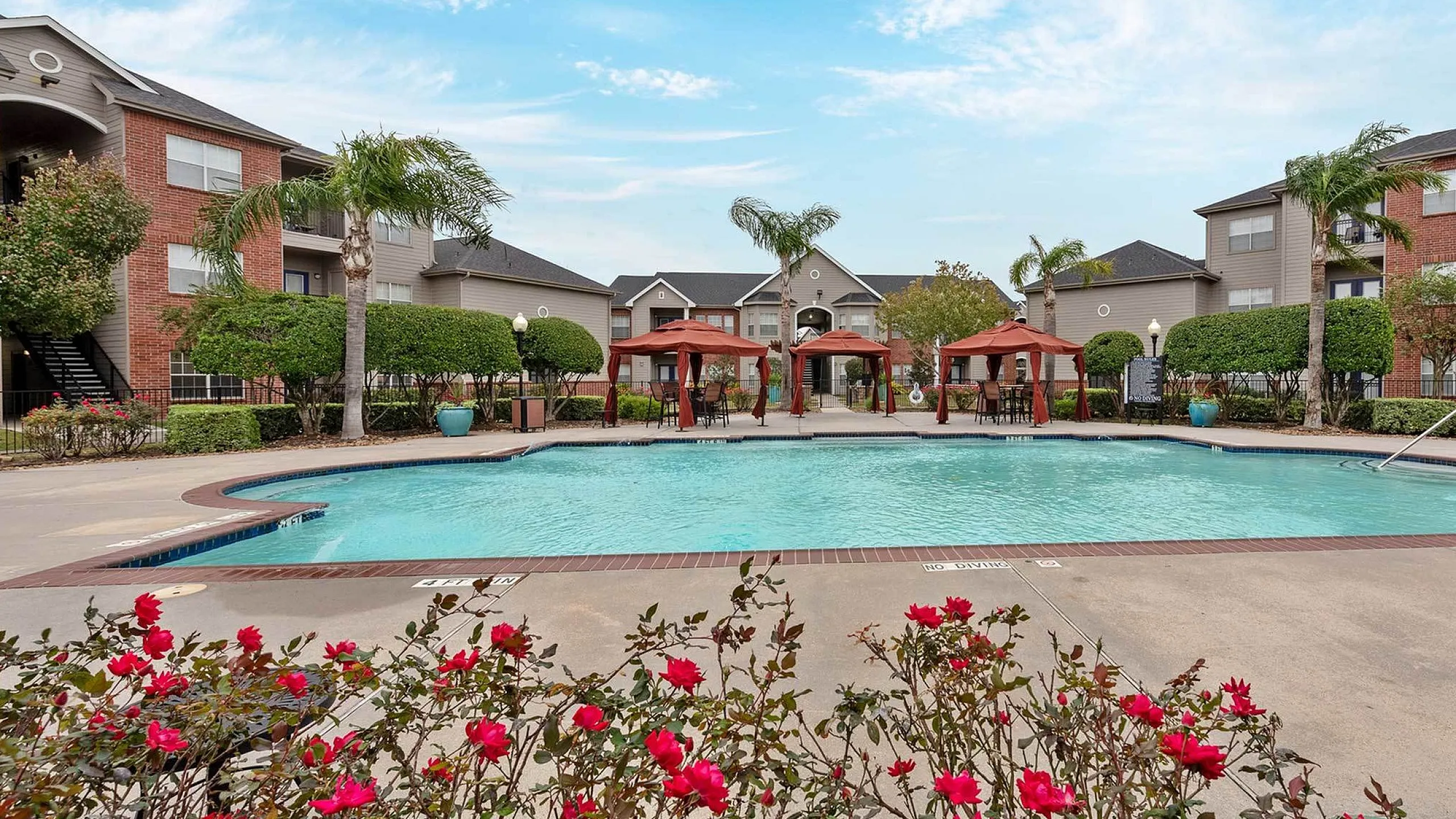 Inviting apartment complex with a sparkling pool surrounded by lush greenery and vibrant red flowers, perfect for relaxation and leisure.