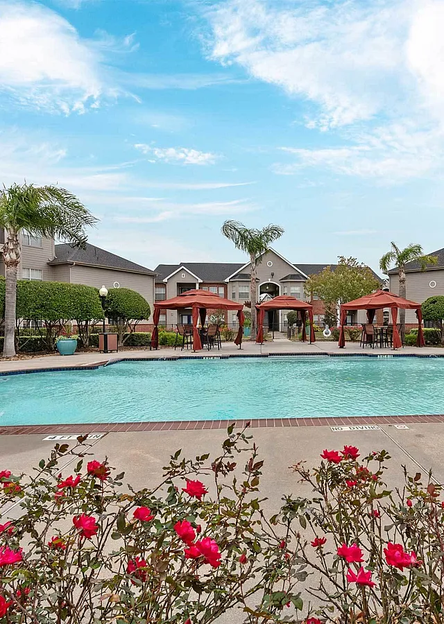 Inviting apartment community with a sparkling swimming pool, lush landscaping, and elegant red-brick buildings under a clear blue sky.