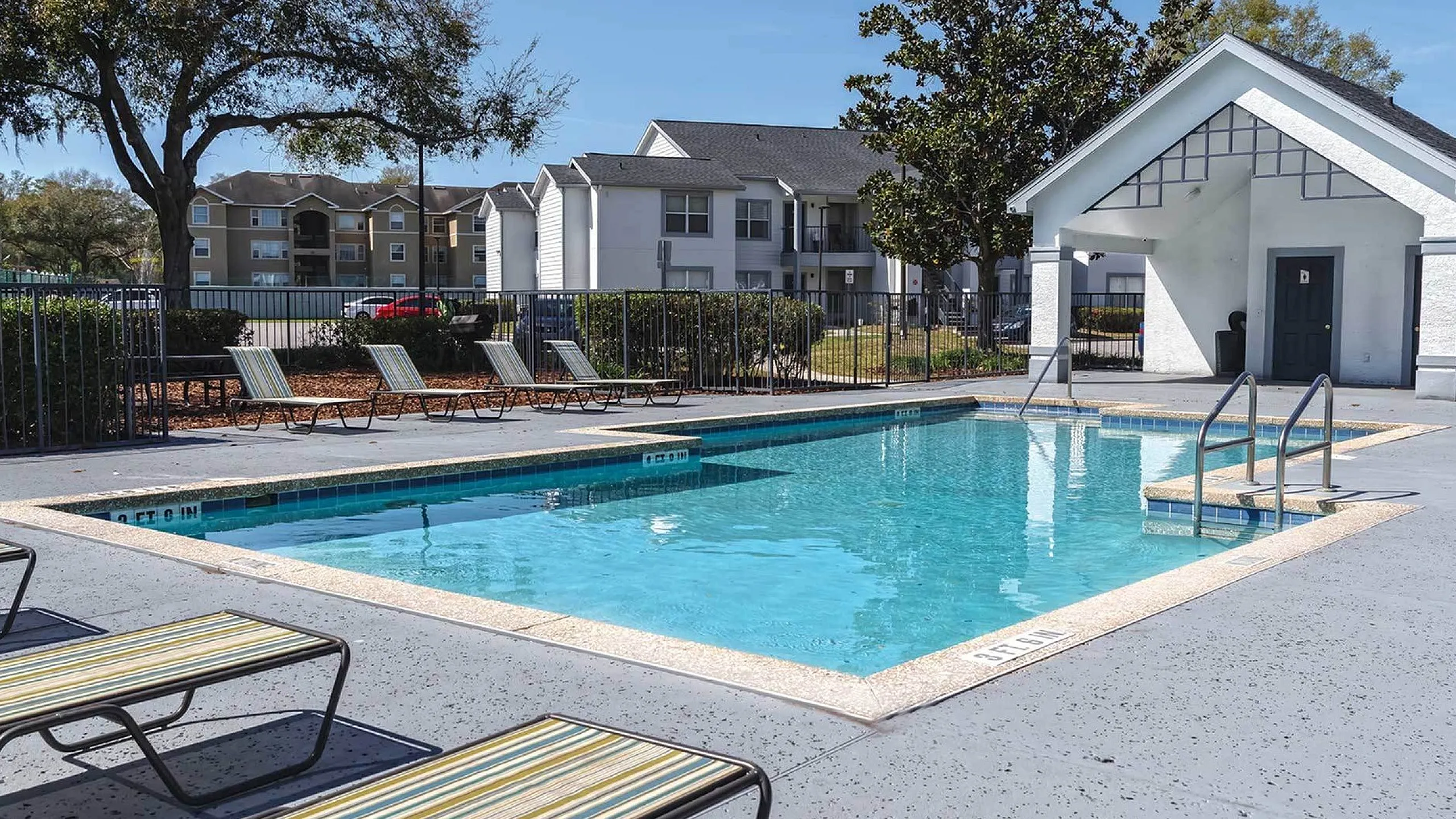 Inviting outdoor swimming pool with lounge chairs, surrounded by lush greenery and modern apartment buildings under a clear blue sky.