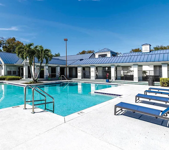 Luxurious apartment complex showcasing a sparkling outdoor pool with sun loungers, lush palm trees, and modern architecture against a bright blue sky.