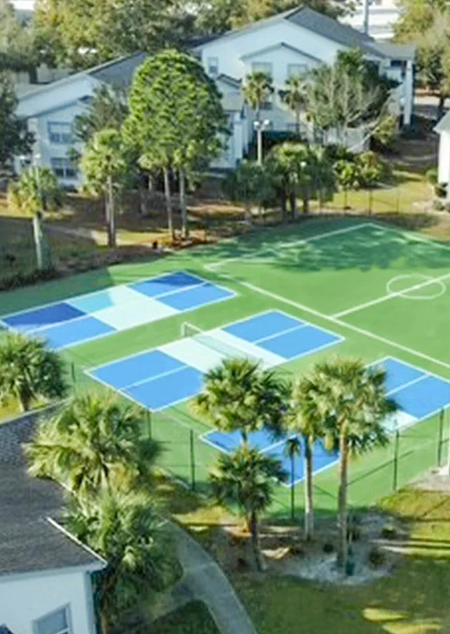 Aerial view of vibrant apartment community featuring a lush green landscape, modern tennis and basketball courts, surrounded by charming white buildings nestled among palm trees.