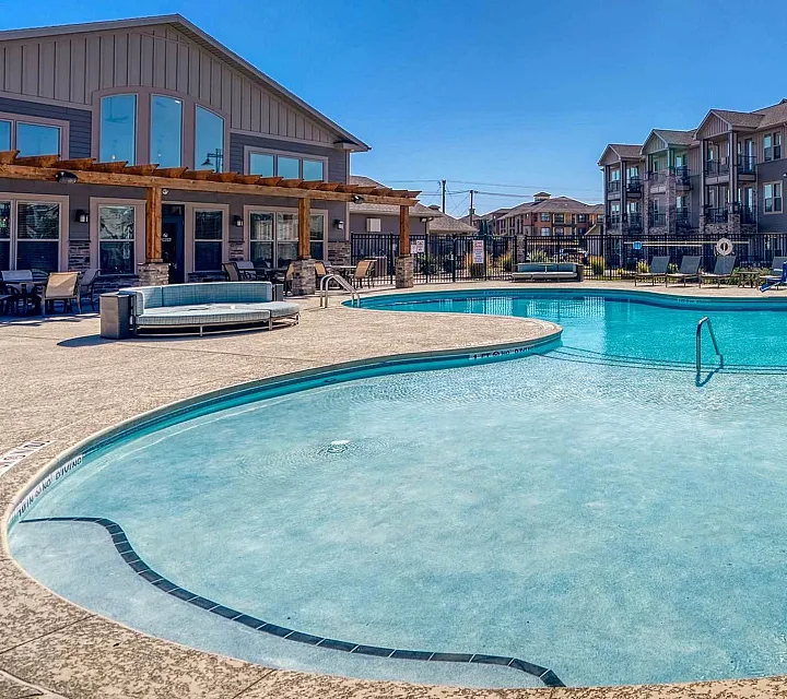 Inviting apartment complex pool area with crystal-clear water, sun loungers, and elegant poolside seating under a bright blue sky.