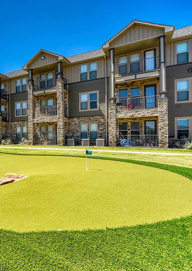 Luxurious apartment complex featuring elegant stonework and private balconies, with a well-maintained putting green under a clear blue sky.