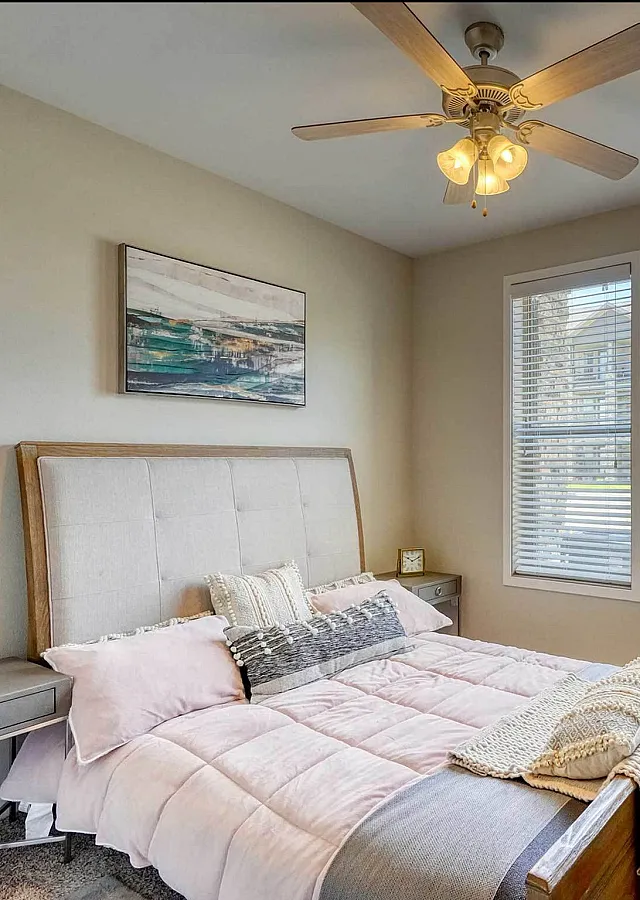 Inviting bedroom with plush bedding, cozy carpet, modern ceiling fan, and natural light streaming through large windows.