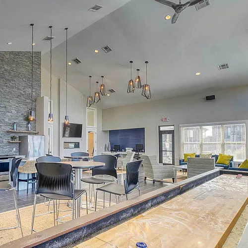 Modern apartment community room featuring a sleek kitchen, stylish seating area, large windows, and a shuffleboard table for entertainment.