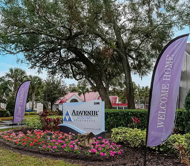Inviting entrance of Advenir Apartments featuring vibrant landscaping, welcoming signage, and lush trees set against a bright blue sky.