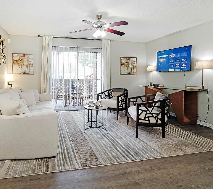 Modern living room with elegant decor, featuring a cozy white sofa, stylish armchairs, and wall-mounted TV. Bright natural light filters through large sliding glass doors, enhancing the chic ambiance.