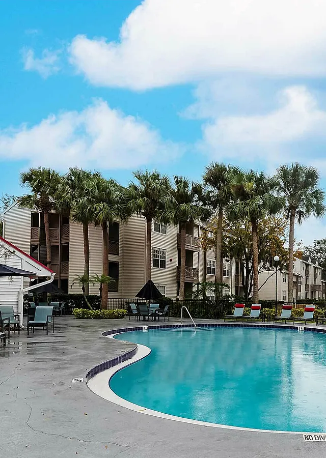 Inviting outdoor pool area at a modern apartment complex featuring lush palm trees, comfortable lounge chairs, and vibrant umbrellas under a bright blue sky.