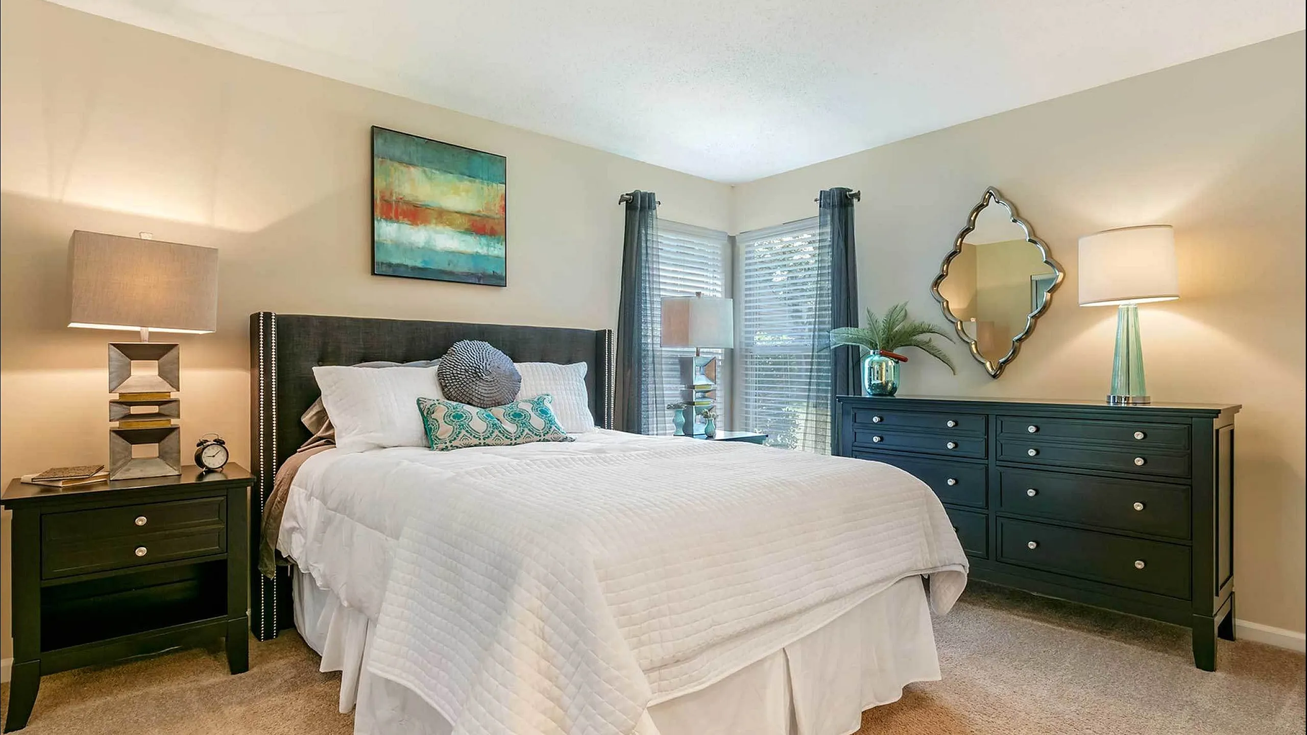 Elegant bedroom with plush carpet, a cozy bed with white linens, stylish nightstands, and a contemporary dresser, highlighted by modern lamps and artistic decor.