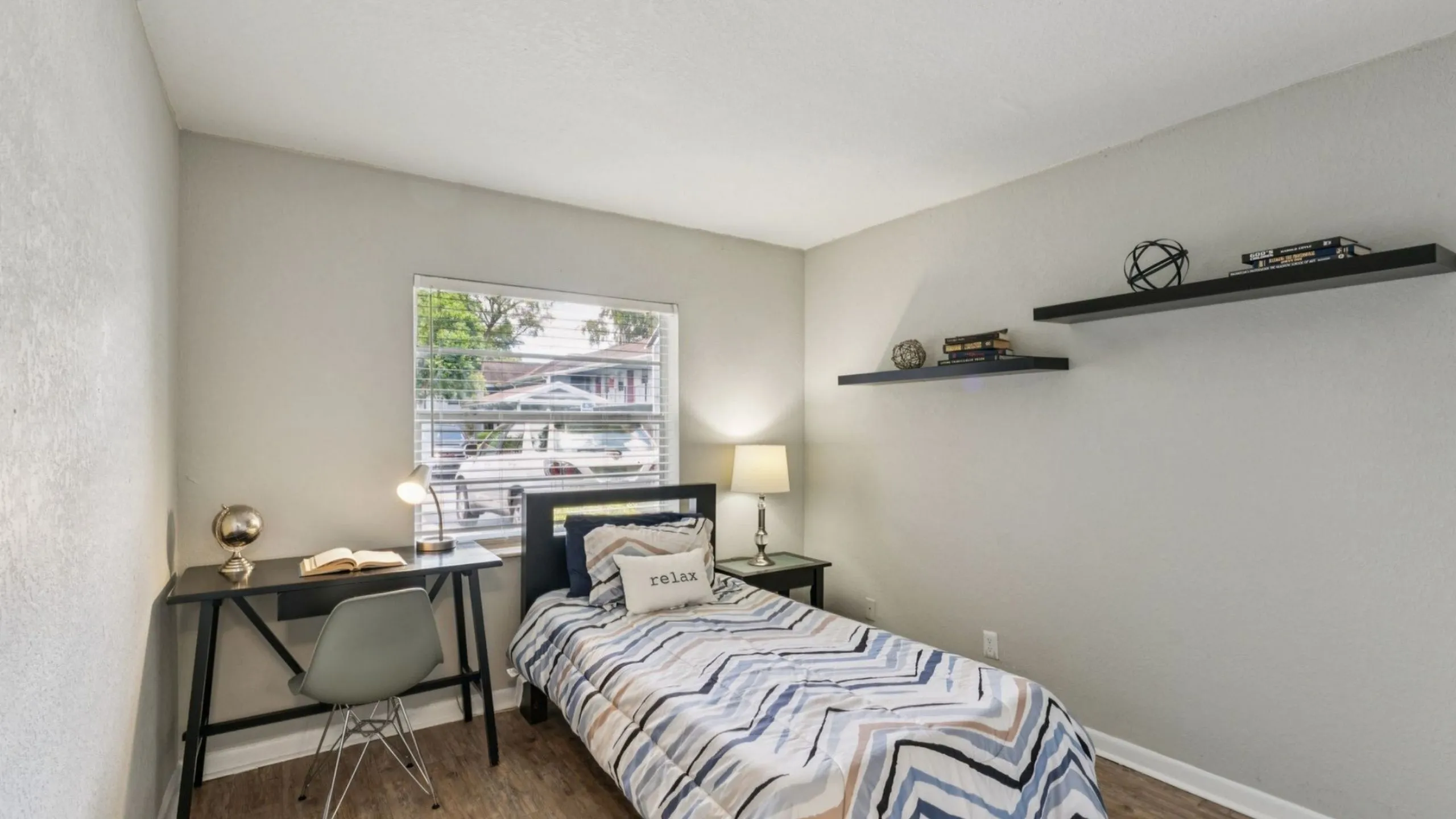 Charming bedroom with a cozy bed, sleek study desk, and elegant wall shelves, filled with natural light from a large window.