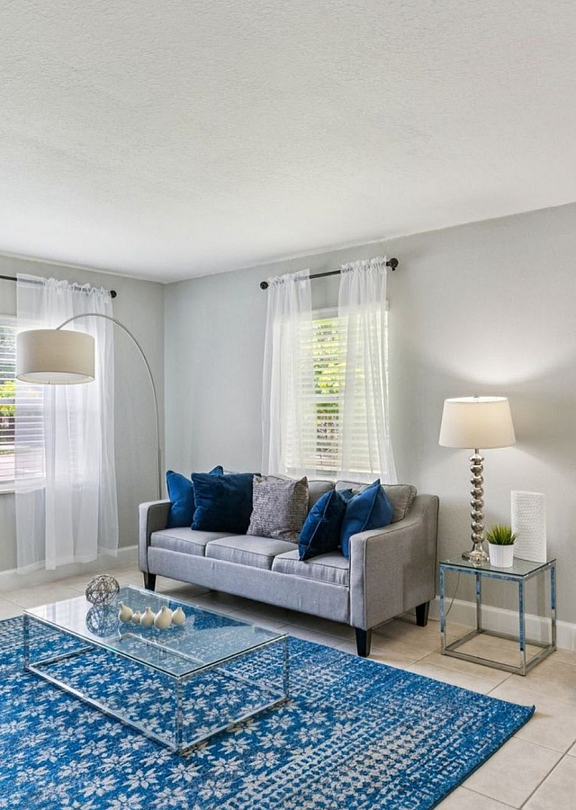 Bright and inviting living room with modern decor, featuring a plush grey sofa, elegant glass coffee table, and artistic wall painting, enhanced by natural light from large windows and sheer curtains.