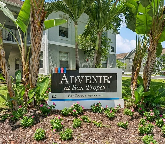 Vibrant garden landscape with a welcoming sign for Advenir at San Tropez, surrounded by lush greenery and tropical plants against a backdrop of modern apartment buildings.