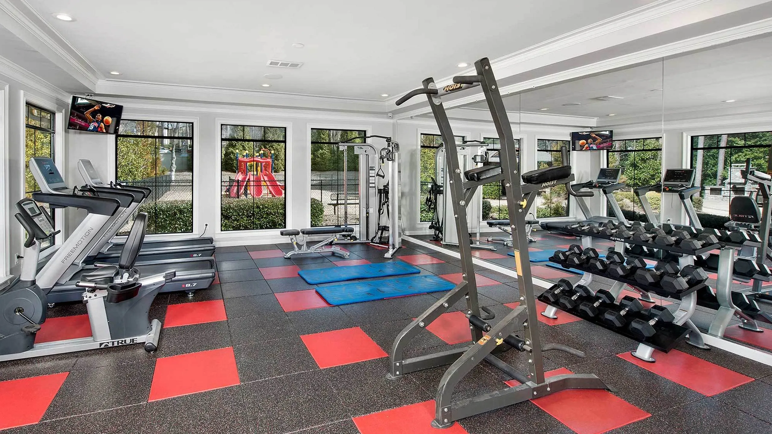 Bright and modern gym with state-of-the-art equipment, including treadmills and free weights, overlooking a vibrant outdoor playground.