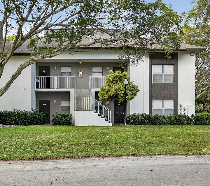 Charming two-story apartment building surrounded by lush greenery and mature trees, showcasing a welcoming exterior with a neatly landscaped lawn.