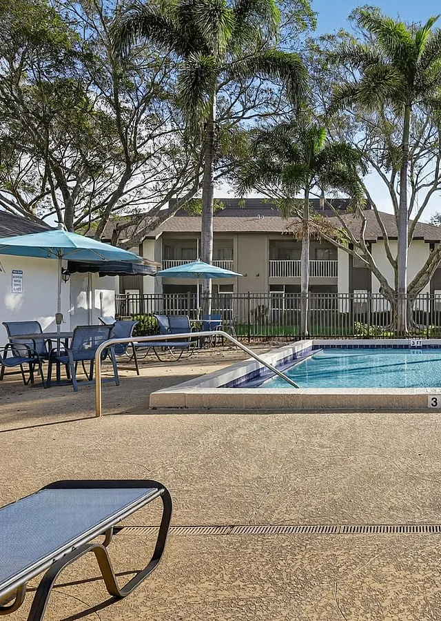 Inviting apartment community pool area surrounded by lush palm trees, featuring comfortable lounge chairs and shaded tables for relaxation.