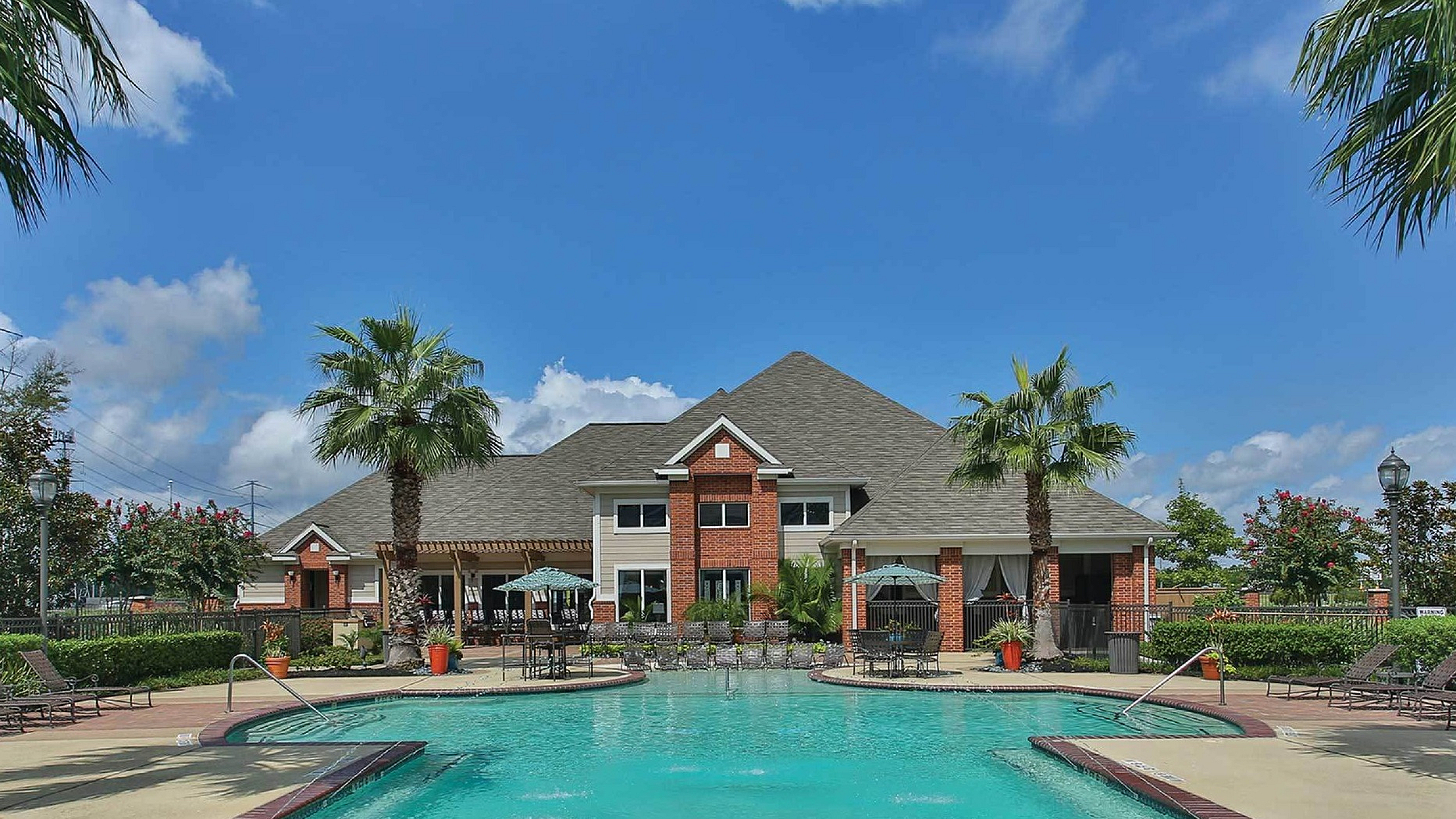 Luxurious apartment complex with a sparkling pool, surrounded by palm trees and lush greenery, under a vibrant blue sky.
