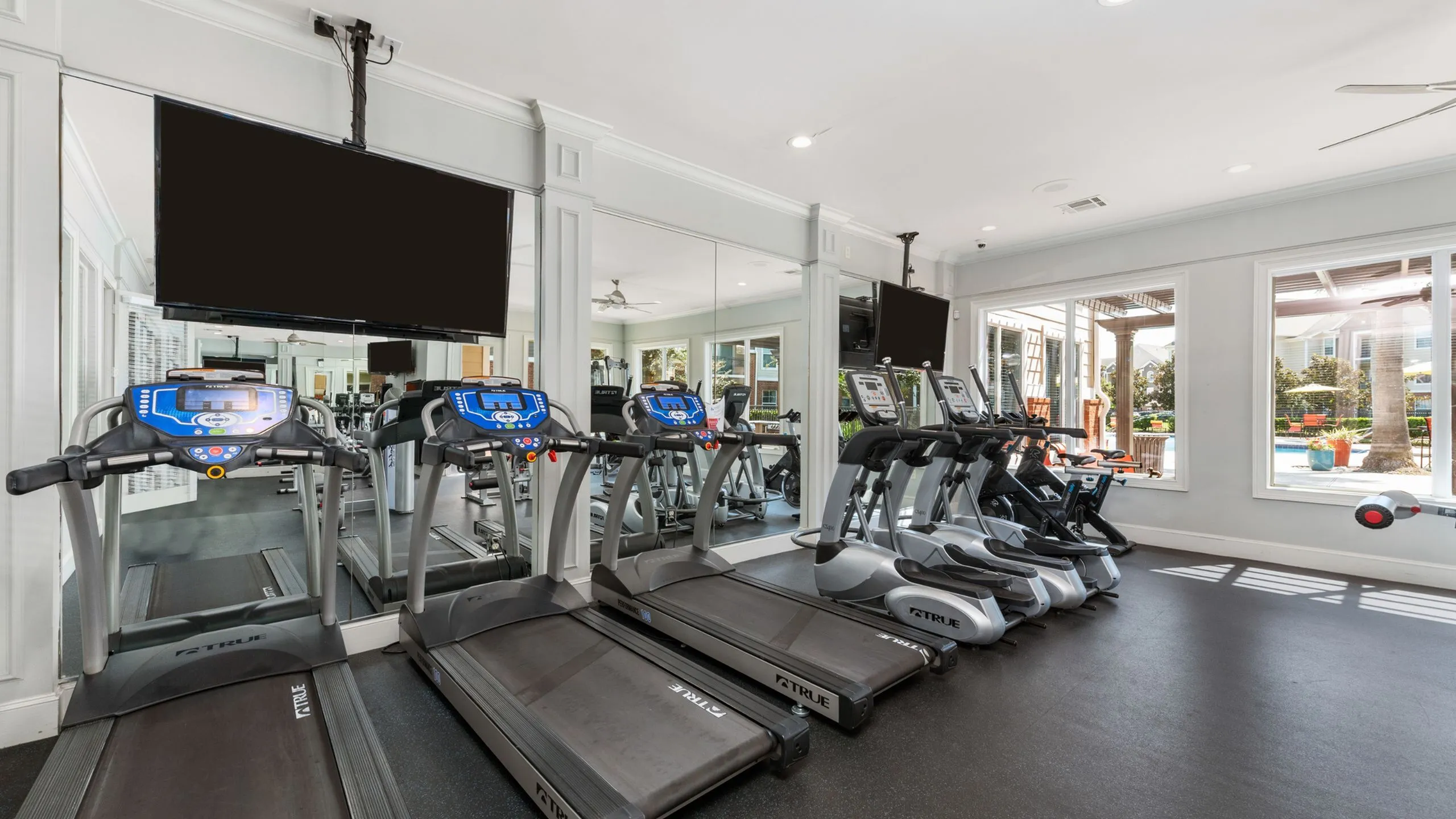Modern fitness center with multiple treadmills, large mirrors, and natural light from expansive windows showcasing a pool view.