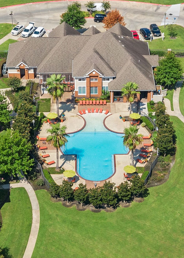 Aerial view of a luxurious apartment complex showcasing a resort-style pool with lush landscaping, surrounded by modern buildings and expansive green spaces.