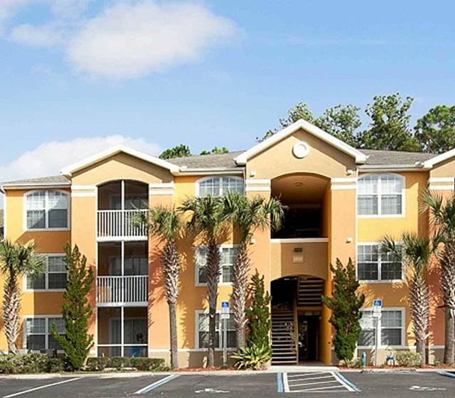 Charming three-story apartment complex with bright orange exterior, lush palm trees, and a welcoming parking area under a clear blue sky.