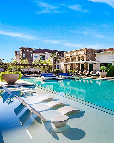Luxurious apartment complex with a stunning outdoor pool, stylish lounge chairs, and vibrant greenery under a clear blue sky.