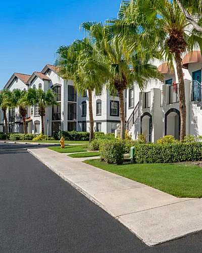Charming apartment complex with modern architecture, surrounded by lush greenery and palm trees, under a bright blue sky.