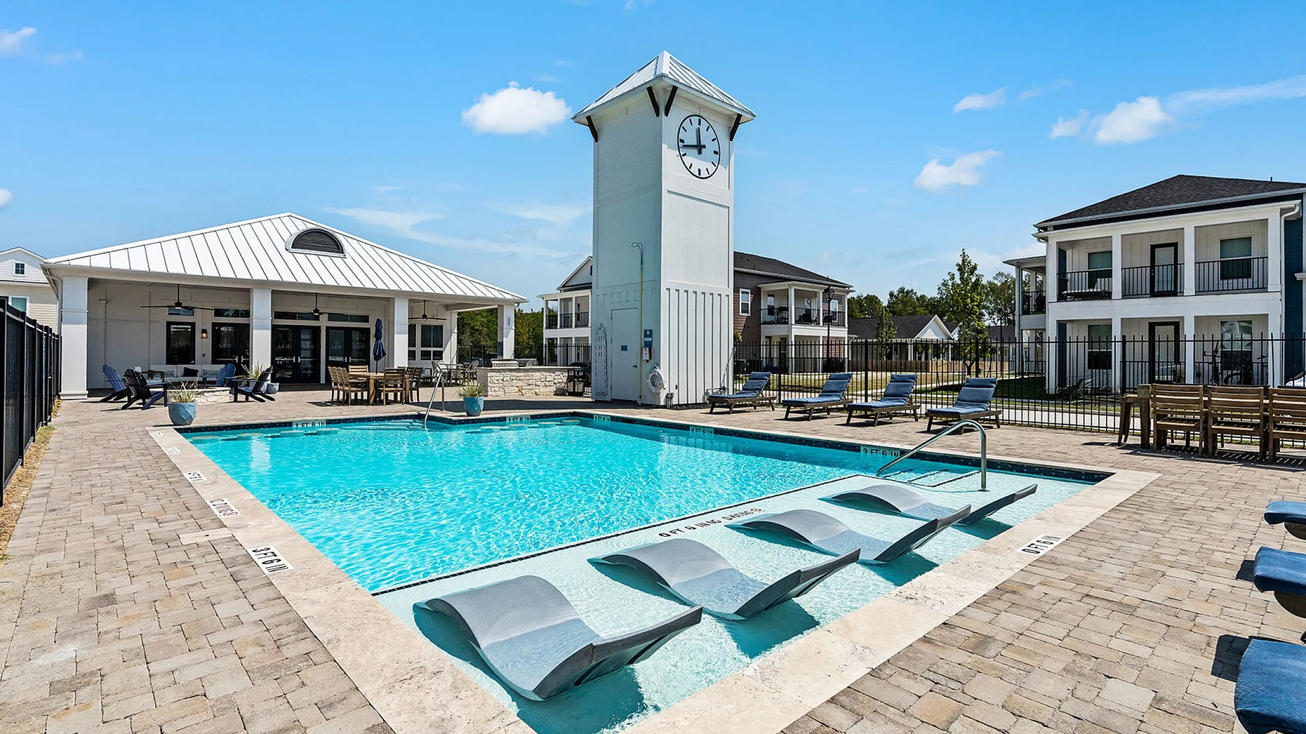 Luxurious apartment community with a sparkling pool, elegant clock tower, and inviting lounge chairs under a bright blue sky.