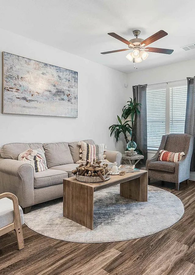 Elegant living room with modern decor, featuring a cozy beige sofa, stylish armchairs, and a wooden coffee table on sleek wooden flooring. Natural light streams through large windows framed by gray curtains.
