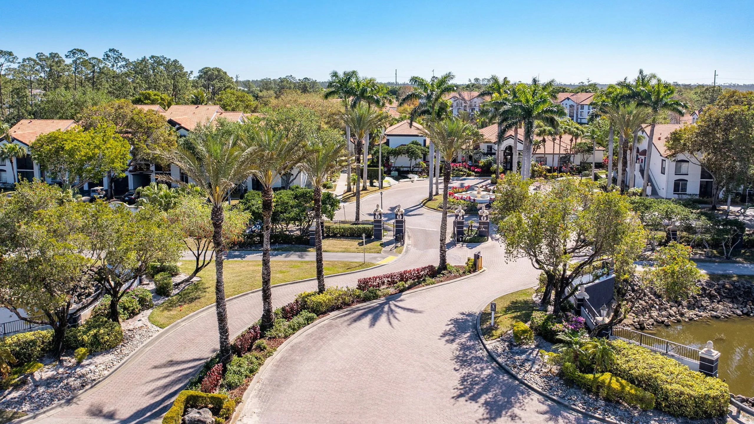 Luxurious apartment complex with lush landscaping, palm-lined entrance, and beautiful water features under a clear blue sky.
