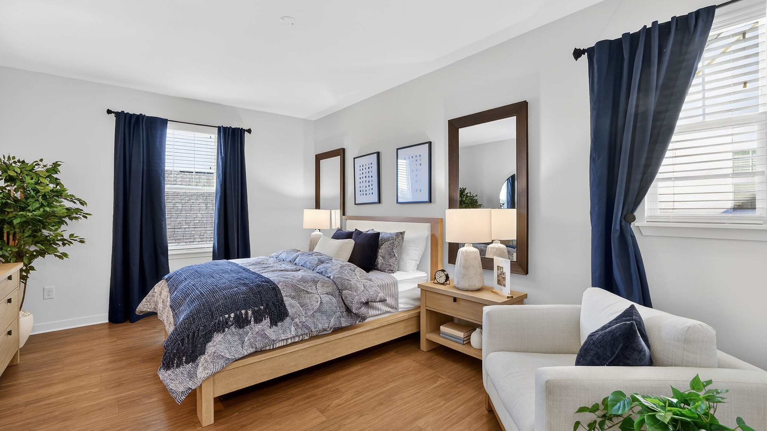 Elegant bedroom with modern decor, featuring a cozy bed with plush pillows, natural wood flooring, and ambient lighting, complemented by deep blue curtains and lush green plants.
