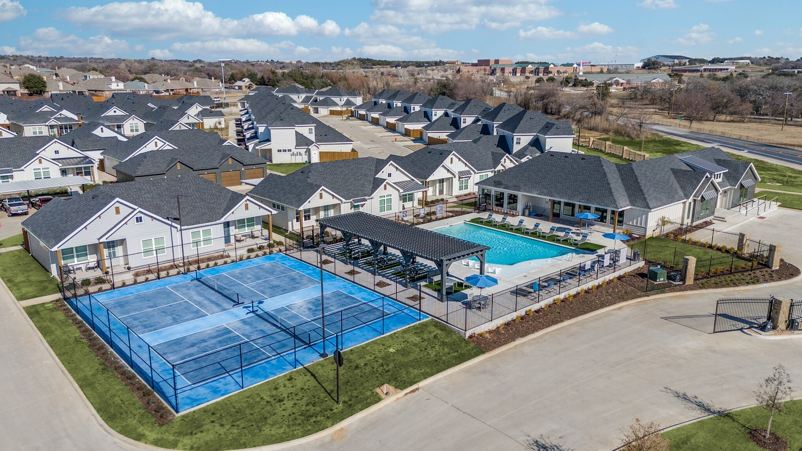 Aerial view of a vibrant residential community featuring modern homes, a refreshing swimming pool, and an expansive tennis court under a clear blue sky.