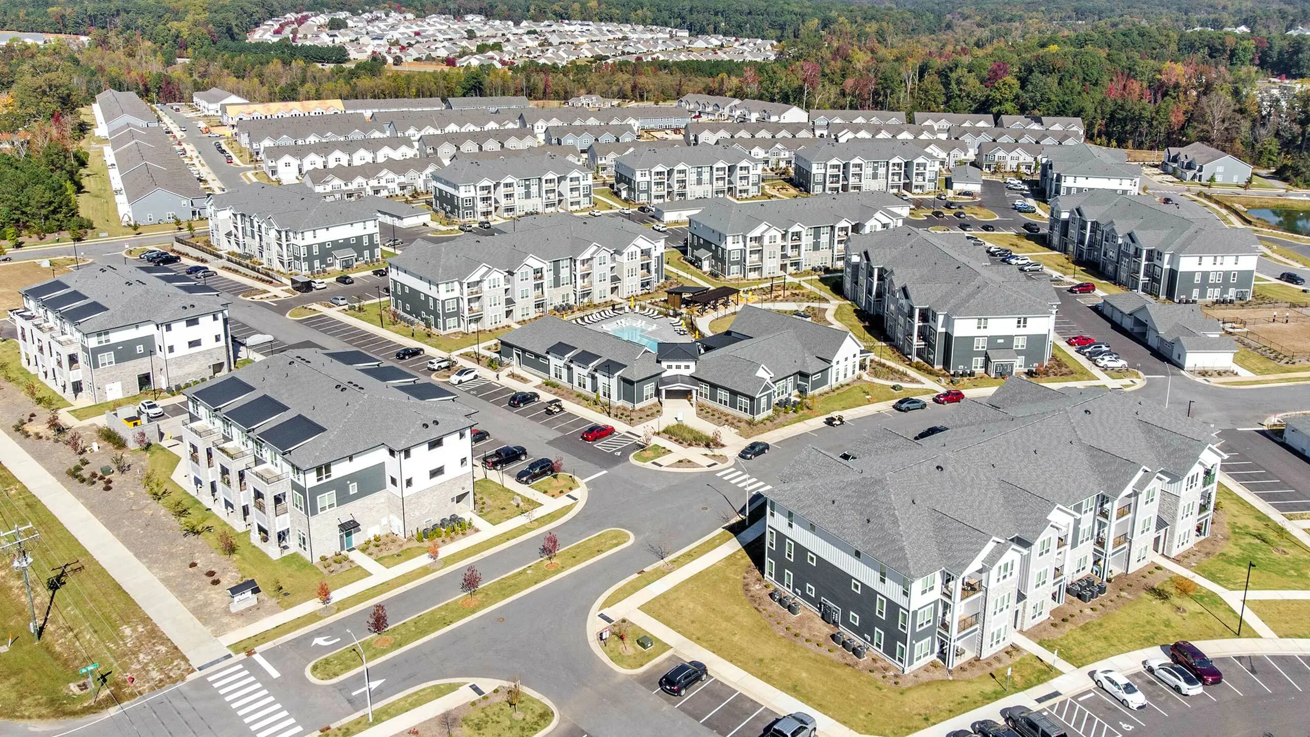 Vibrant aerial view of a modern apartment community with spacious buildings, green spaces, and a central swimming pool, surrounded by scenic landscapes.