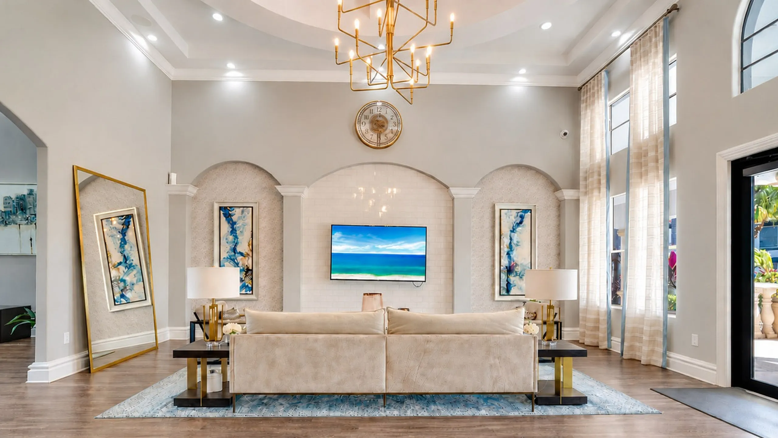 Luxurious and expansive living room featuring elegant decor, plush seating, large windows with sheer curtains, and contemporary wall art complement the chic chandelier lighting.