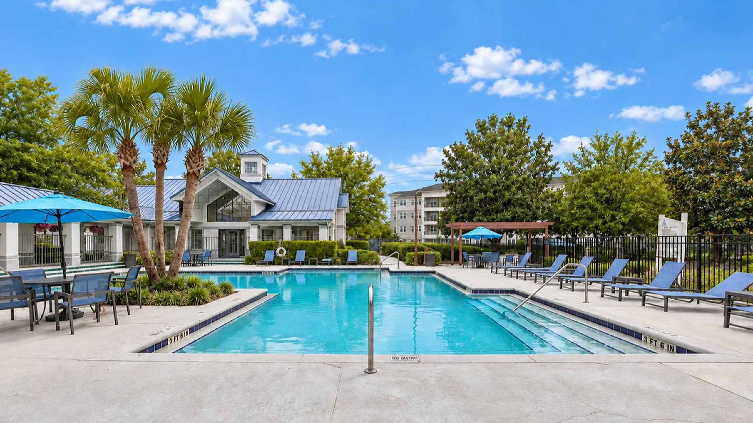 Luxurious outdoor pool area surrounded by lush greenery and modern lounge chairs, featuring a stylish clubhouse with a blue roof and inviting outdoor seating.