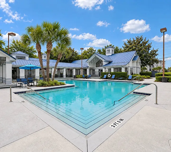 Luxurious apartment pool area with crystal-clear water, elegant lounging chairs, and vibrant blue umbrellas against a backdrop of lush greenery and modern architecture.