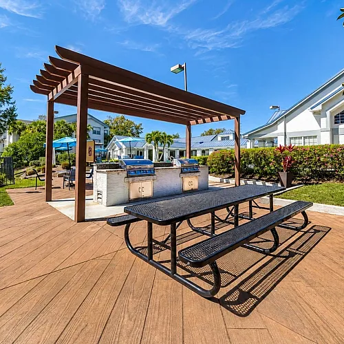 Charming outdoor BBQ area with modern grills and picnic tables set under a wooden pergola, surrounded by lush greenery and residential buildings.