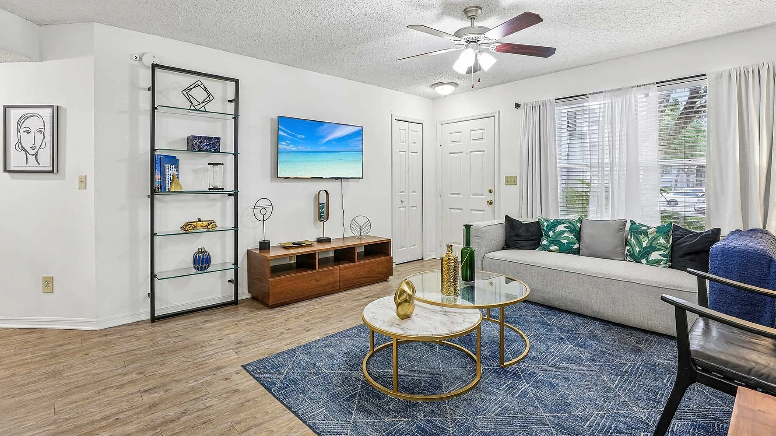 Elegant living room with modern decor, featuring a cozy gray sofa adorned with tropical print cushions, a sleek glass coffee table on a stylish blue rug, and a wall-mounted TV for ultimate relaxation.