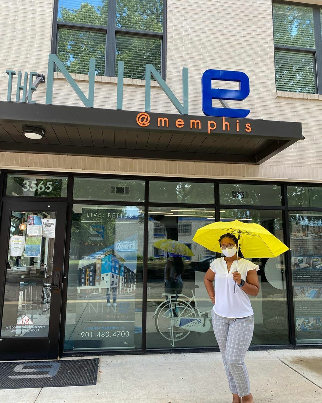 Inviting entrance of The Nine at Memphis, featuring modern design and vibrant signage. A person with a bright yellow umbrella adds a lively touch to the scene.