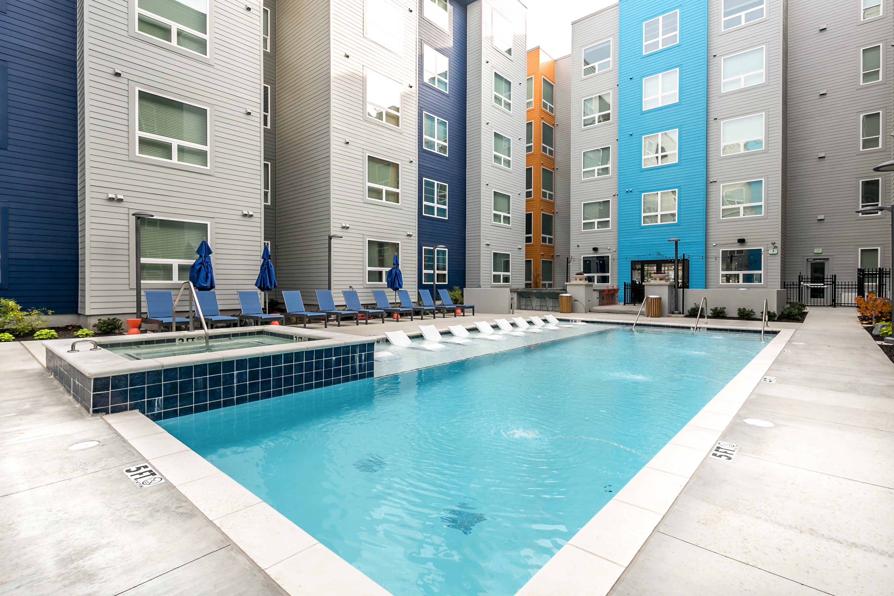 Modern apartment complex with a pristine swimming pool, surrounded by stylish lounge chairs and vibrant buildings in refreshing colors.