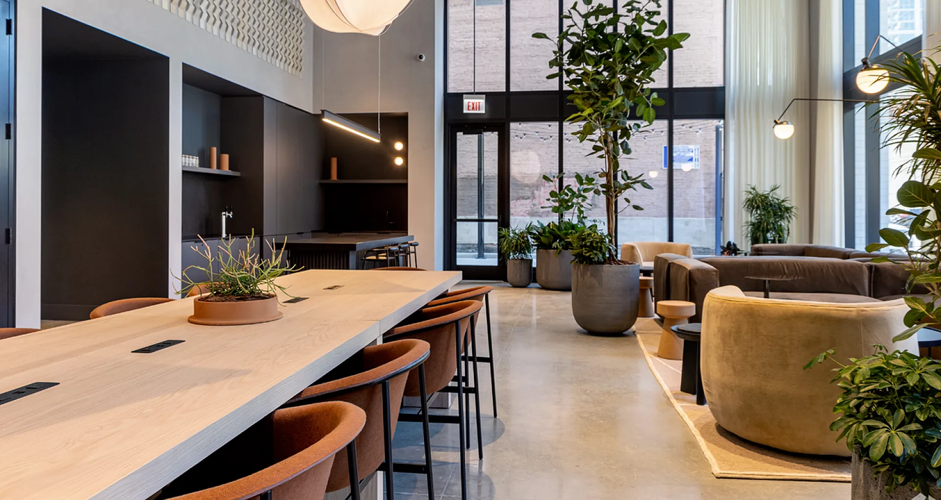 Contemporary apartment lounge with elegant lighting, wooden community table, chic seating, and lush greenery.