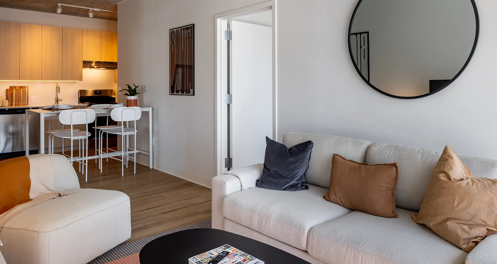 Chic living space boasting a cozy sofa with stylish cushions, modern kitchen with sleek cabinetry, and a minimalist round mirror, perfect for urban living.
