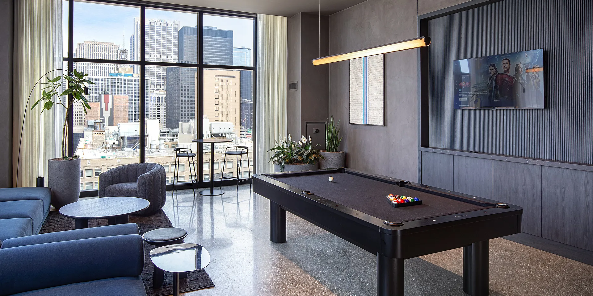Luxurious apartment lounge with a sleek pool table, plush blue seating, and panoramic city views through large windows.