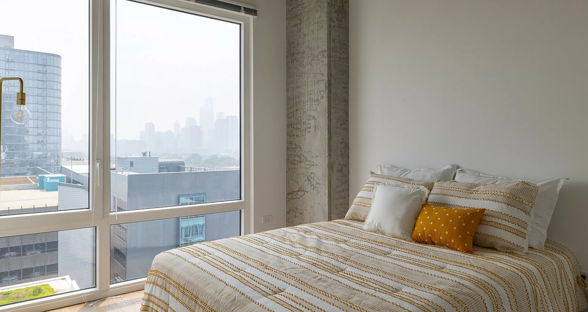 Bright and modern bedroom with large windows offering stunning city views, featuring cozy bedding and a stylish floor lamp.