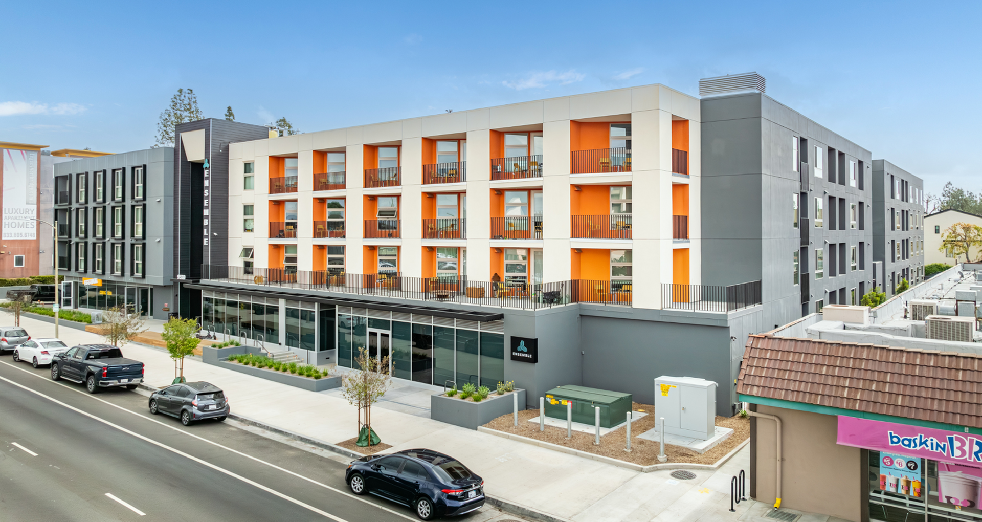 Modern apartment complex with vibrant orange accents and large windows, offering urban living with convenient street-level shops in a lively neighborhood.