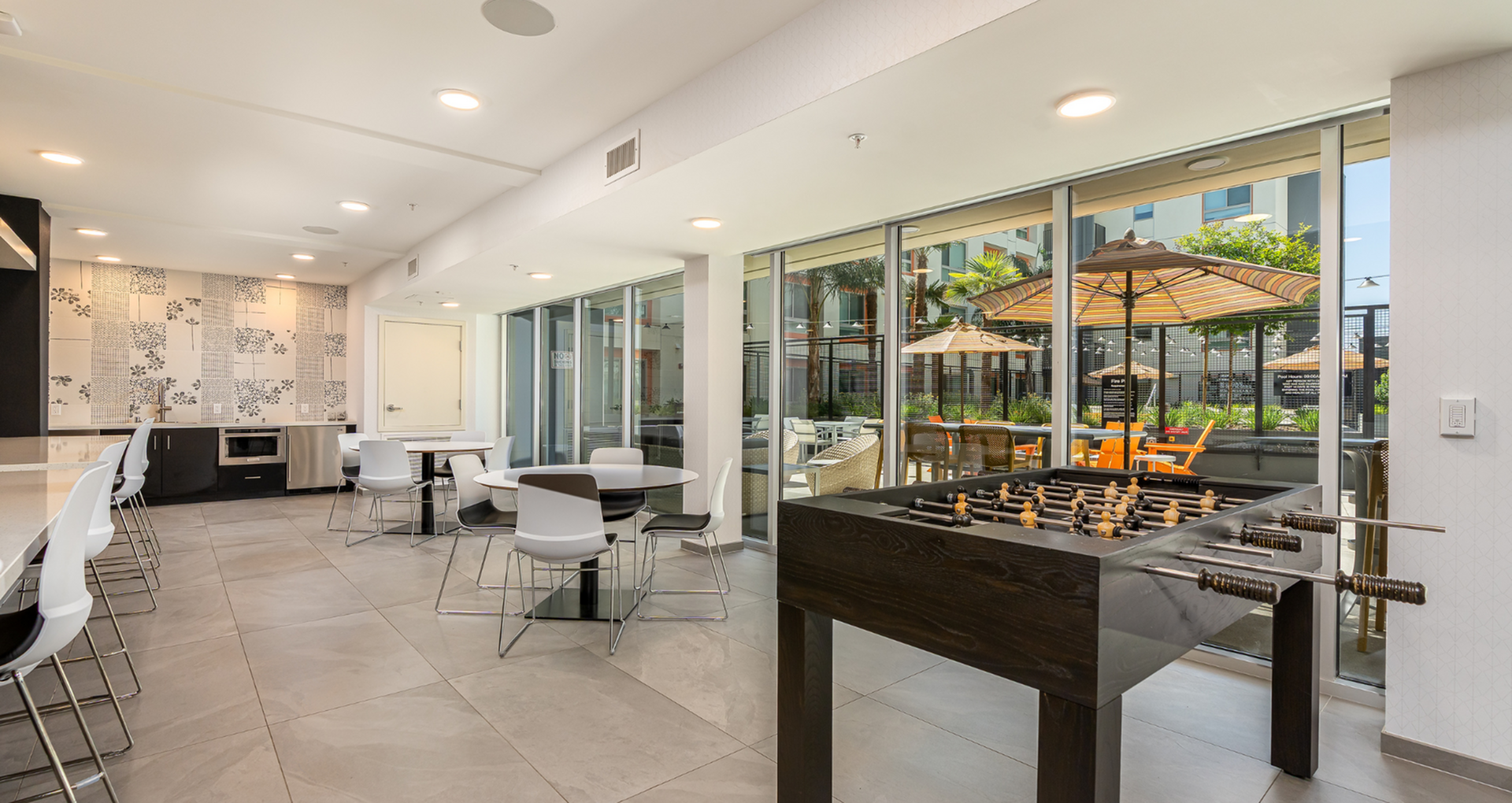 Modern recreation room with sleek dining tables, stylish kitchen area, and large glass doors opening to a vibrant outdoor patio with umbrellas and seating.