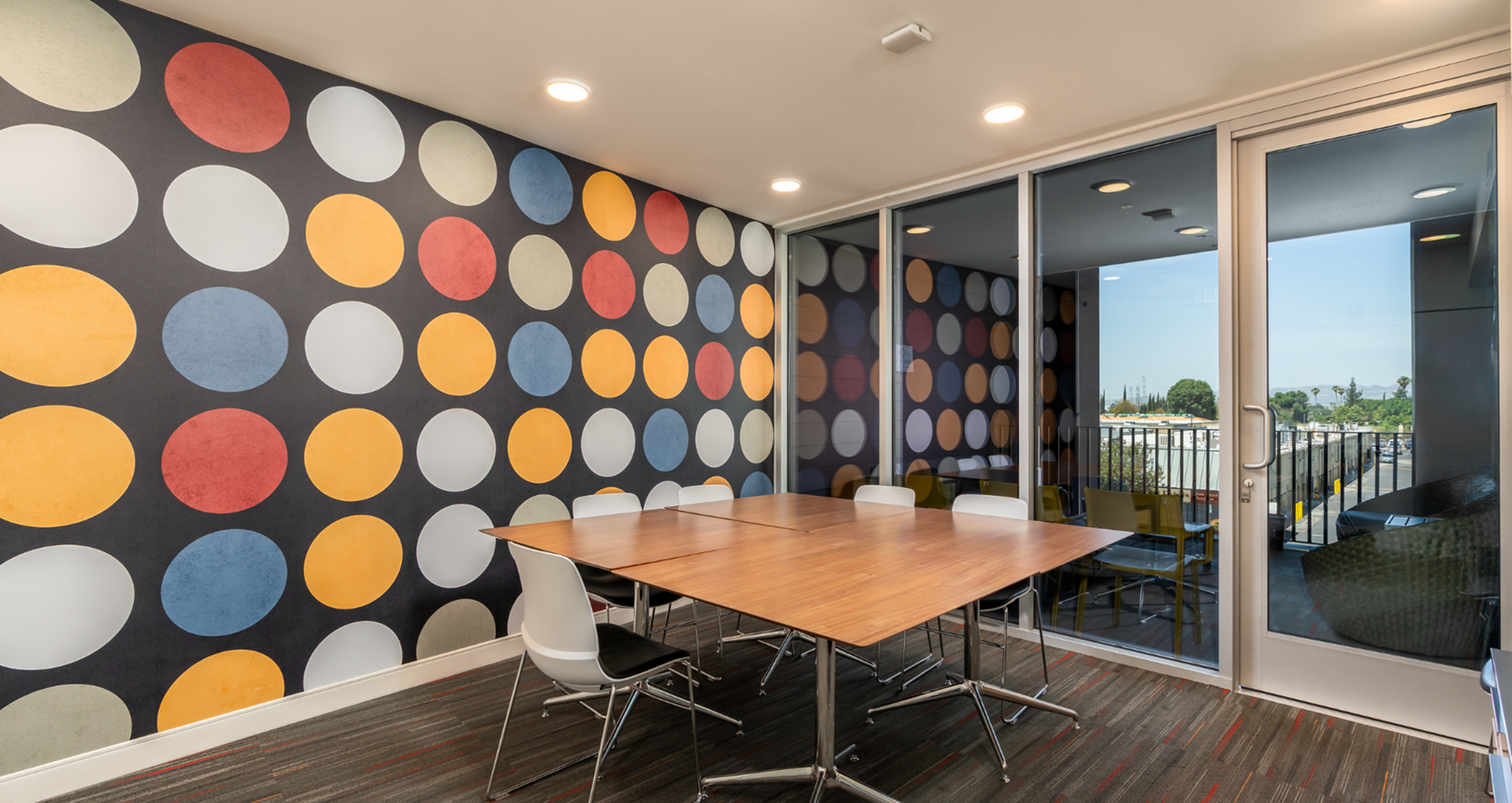 Vibrant meeting room with colorful polka dot wall, sleek wooden table, and stylish chairs, overlooking a scenic view.
